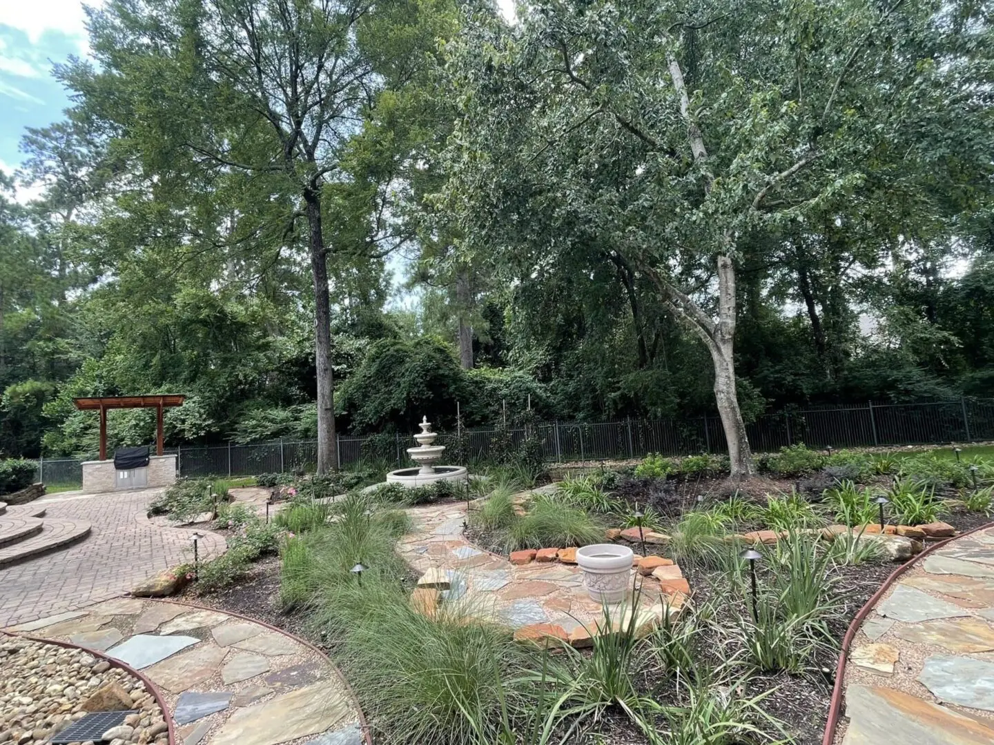 Finished backyard design in The Woodlands with outdoor kitchen pergola, stone fountain, flagstone pathways, and landscaped garden beds.