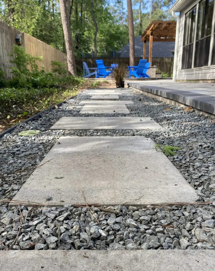 Backyard walkway in The Woodlands, Texas with oversized concrete pavers set in gravel leading to a pergola and seating area with blue Adirondack chairs.