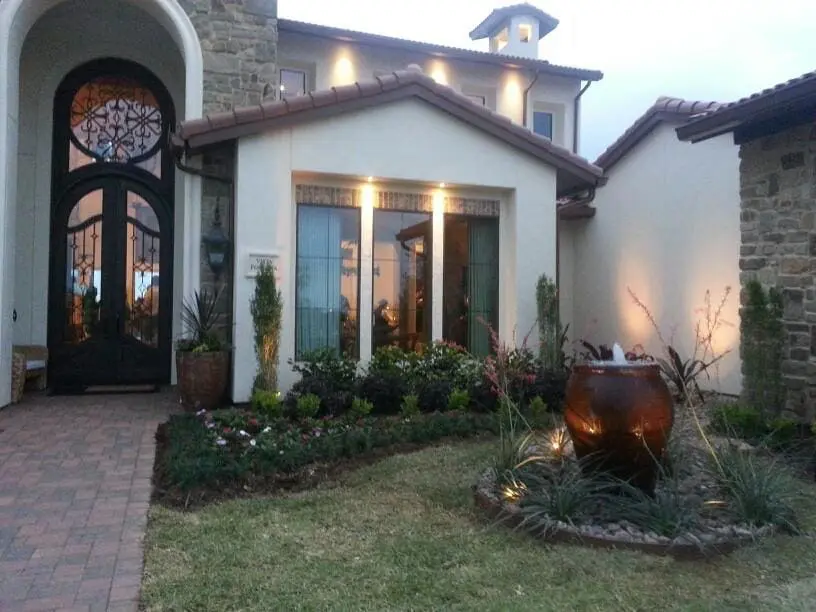 Front entry landscape with illuminated urn fountain and soft lighting at AISD Showcase Home by JM Outdoor Living in The Woodlands, TX.