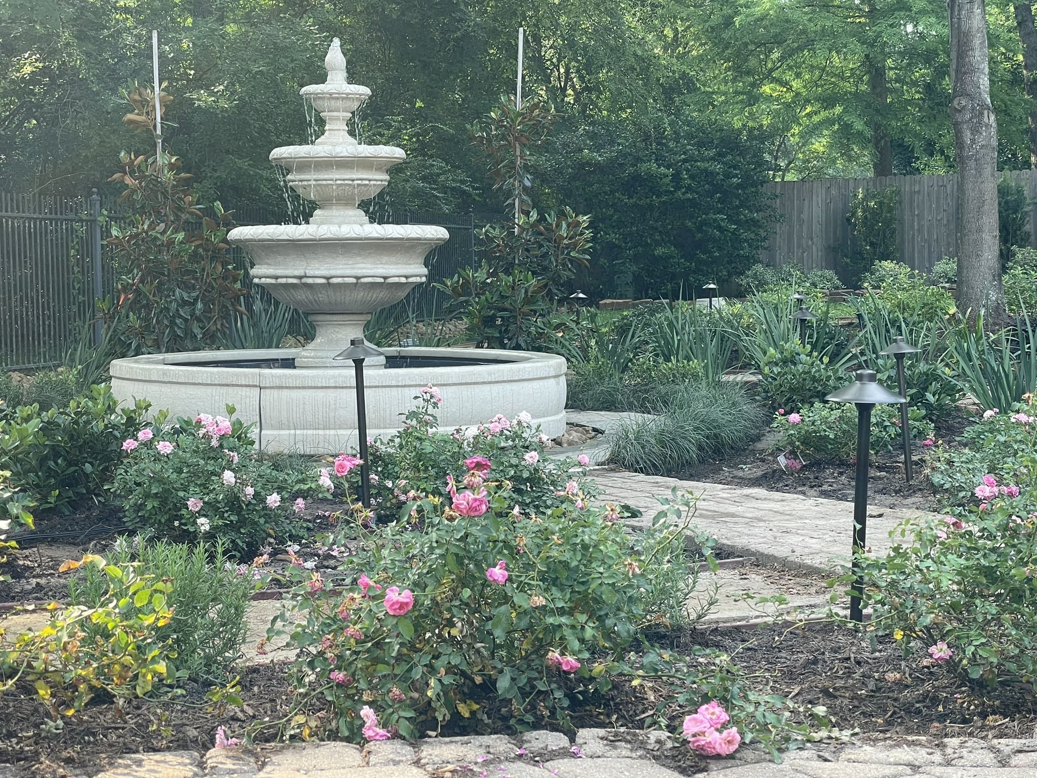 A white tiered fountain in a garden with blooming pink flowers and greenery.