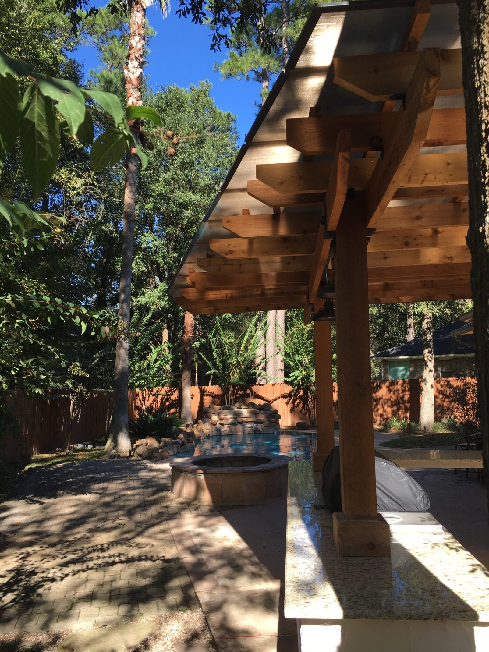 Cedar cantilevered pergola with translucent roof, exposed rafters, and granite outdoor kitchen countertops overlooking pool and waterfall in The Woodlands, Texas.