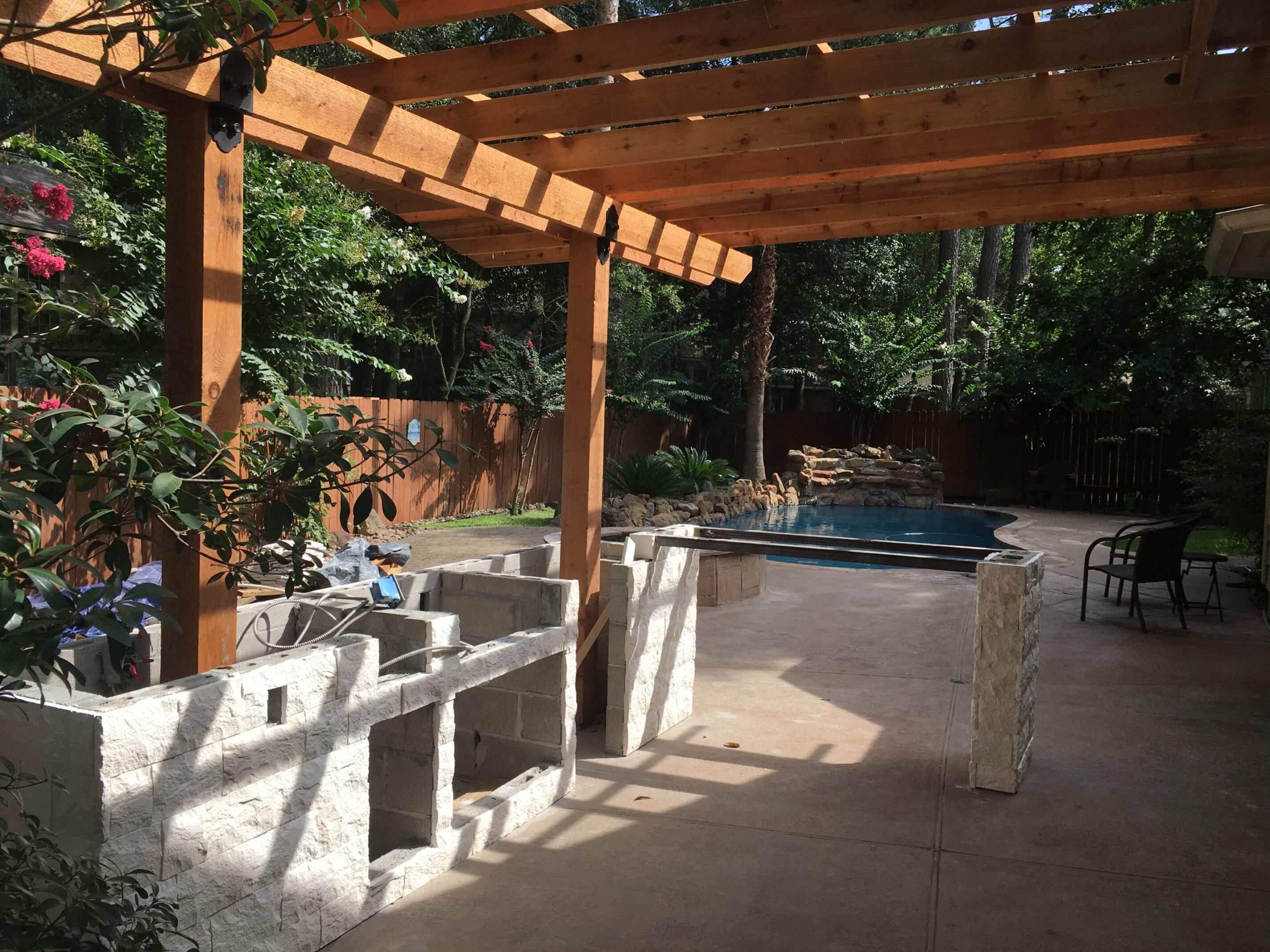 Progress photo of outdoor kitchen with stone veneer under cedar pergola overlooking backyard pool and waterfall in The Woodlands, Texas.