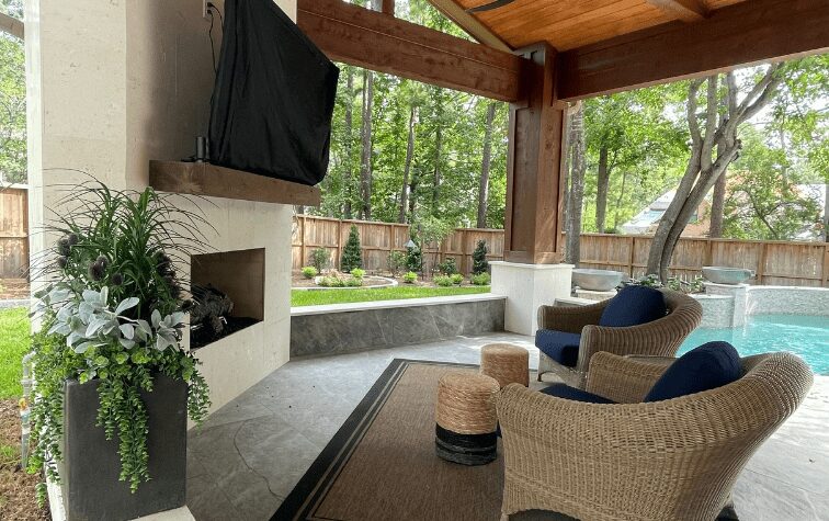 Luxury backyard in The Woodlands, TX with covered patio, stone outdoor fireplace, pool, and lush landscaping by JM Outdoor Living.